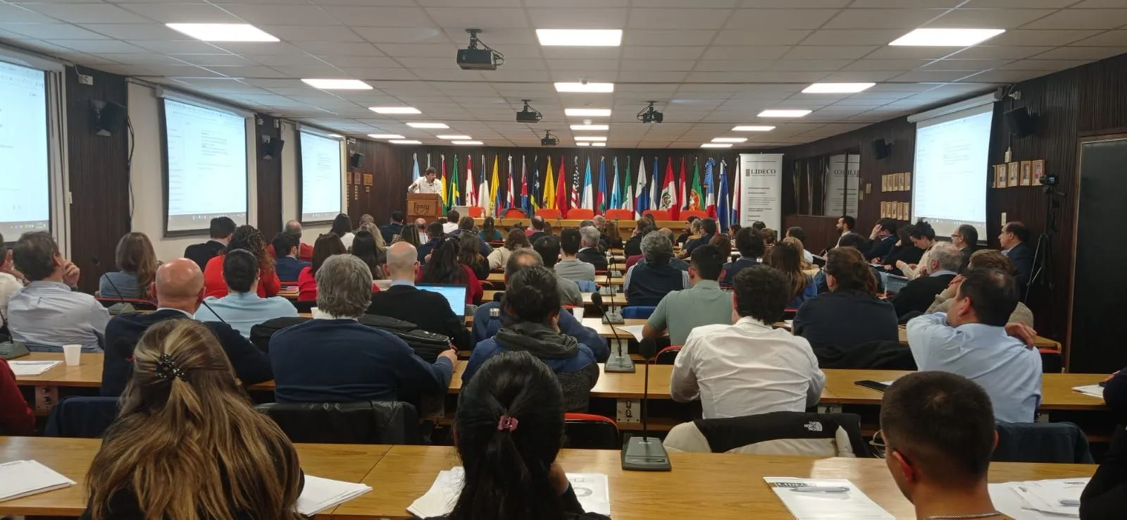 Auditorio lleno con banderas de varios países durante una presentación de Instituto CPE en LIDECO